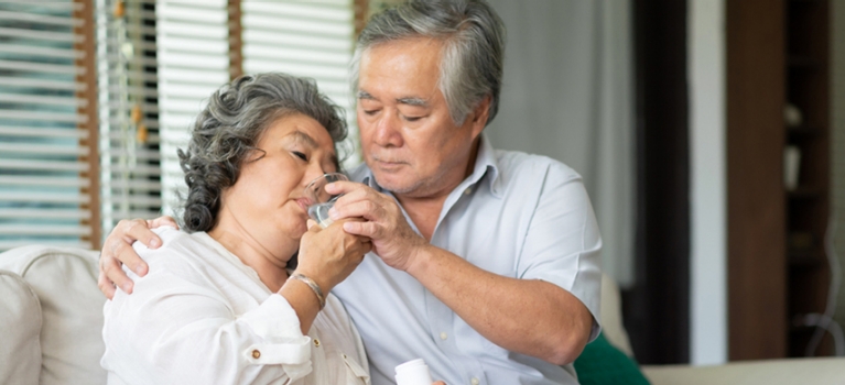 Asian husband helping his sick wife take her medicine while seated on their sofa