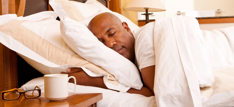 Bald African-American asleep in his bed, with his glasses and coffee mug on his nightstand.