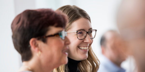 Two women wearing glasses, smiling and engaged in conversation in a bright, professional setting.