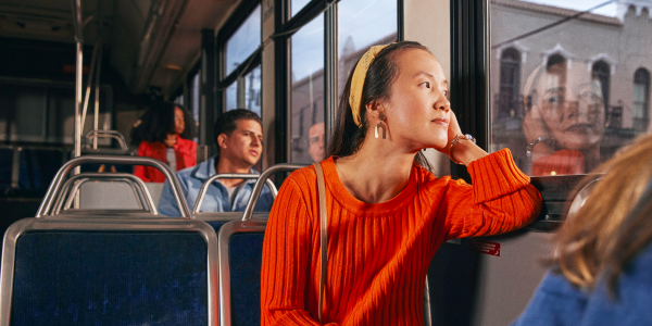 Woman in an orange sweater and yellow headband gazing out of a bus window, with other passengers seated around her.