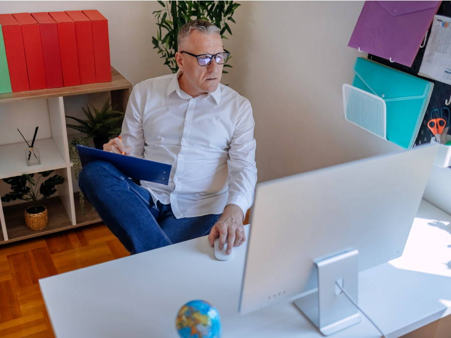 Man in a white shirt working at a desk with a computer, holding a clipboard, in an office.