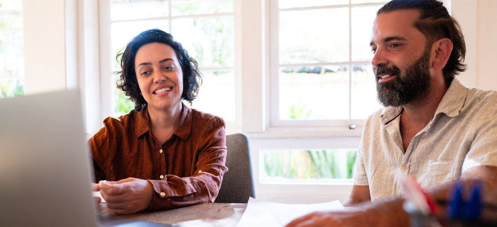 wo colleagues in a bright room smiling and looking at a laptop. The woman has curly short hair and a brown shirt, while the man has a beard and a light shirt.