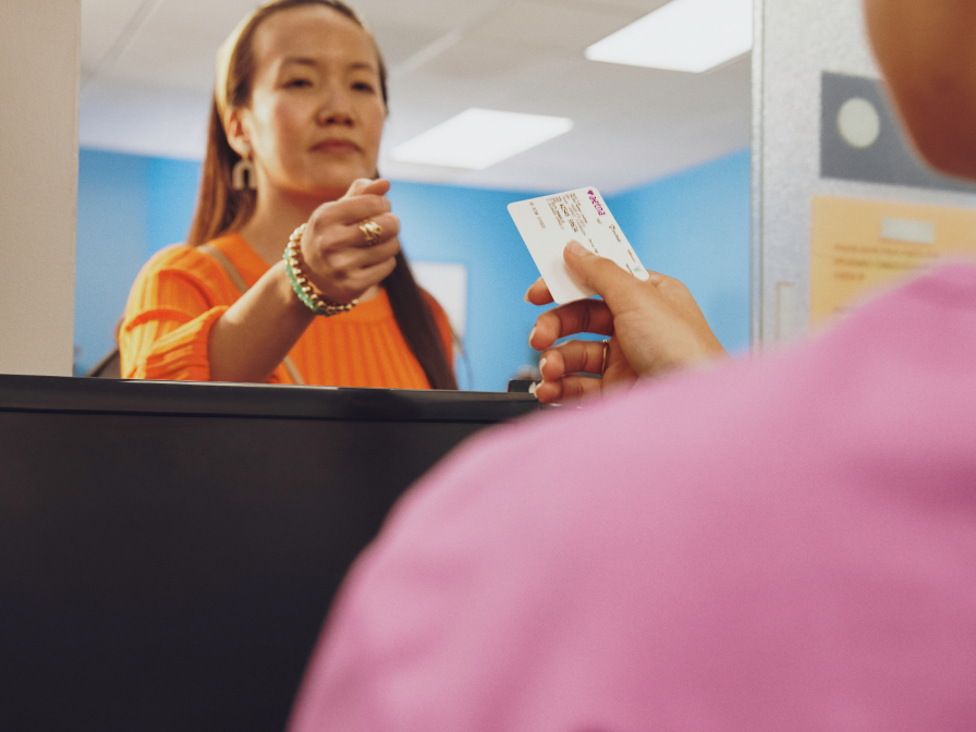 A woman in an orange shirt hands over an Aetna ID card to a person in a pink shirt across a counter.
