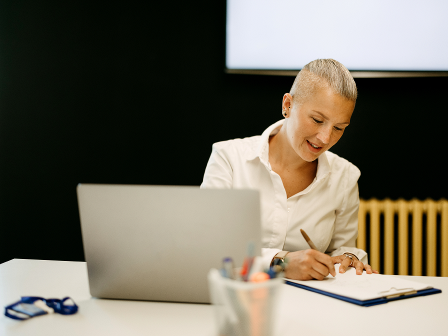 A woman with short blonde hair is sitting at a desk, writing in a notebook with a laptop open in front of her.